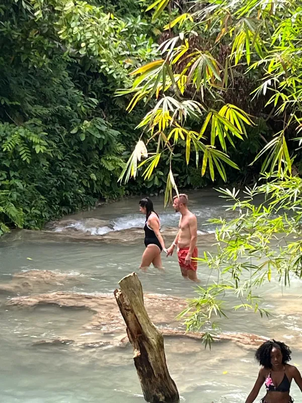 Guests walking across shallow river rocks beneath tropical trees